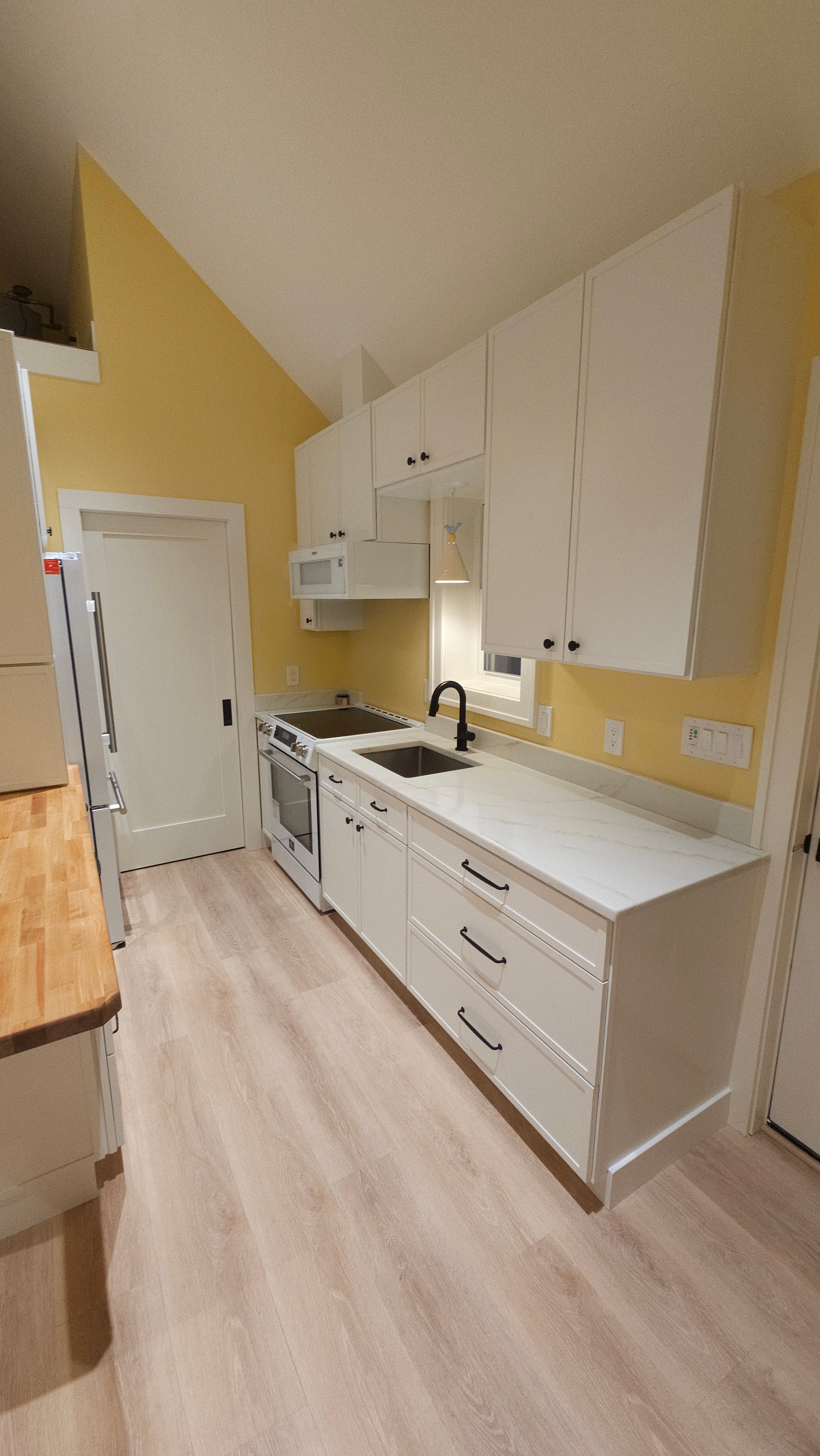 Kitchen cabinet run in the Medway ADU showing white upper and lower cabinets, black hardware, wood countertop edge, and light-toned flooring.