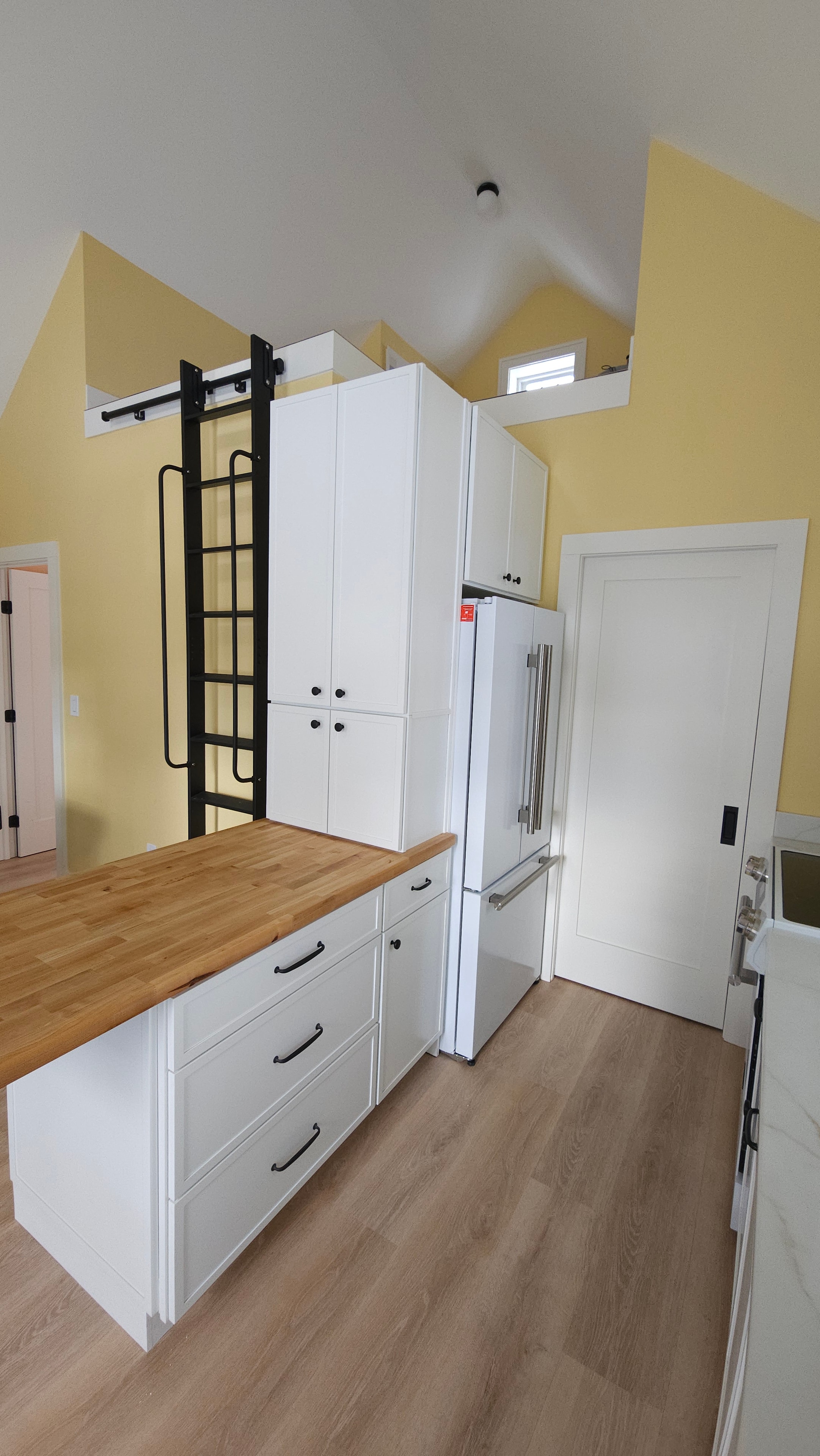 Side view of the Medway ADU kitchen showing tall white cabinetry, the island with wood countertop, loft access ladder, and vaulted ceiling.