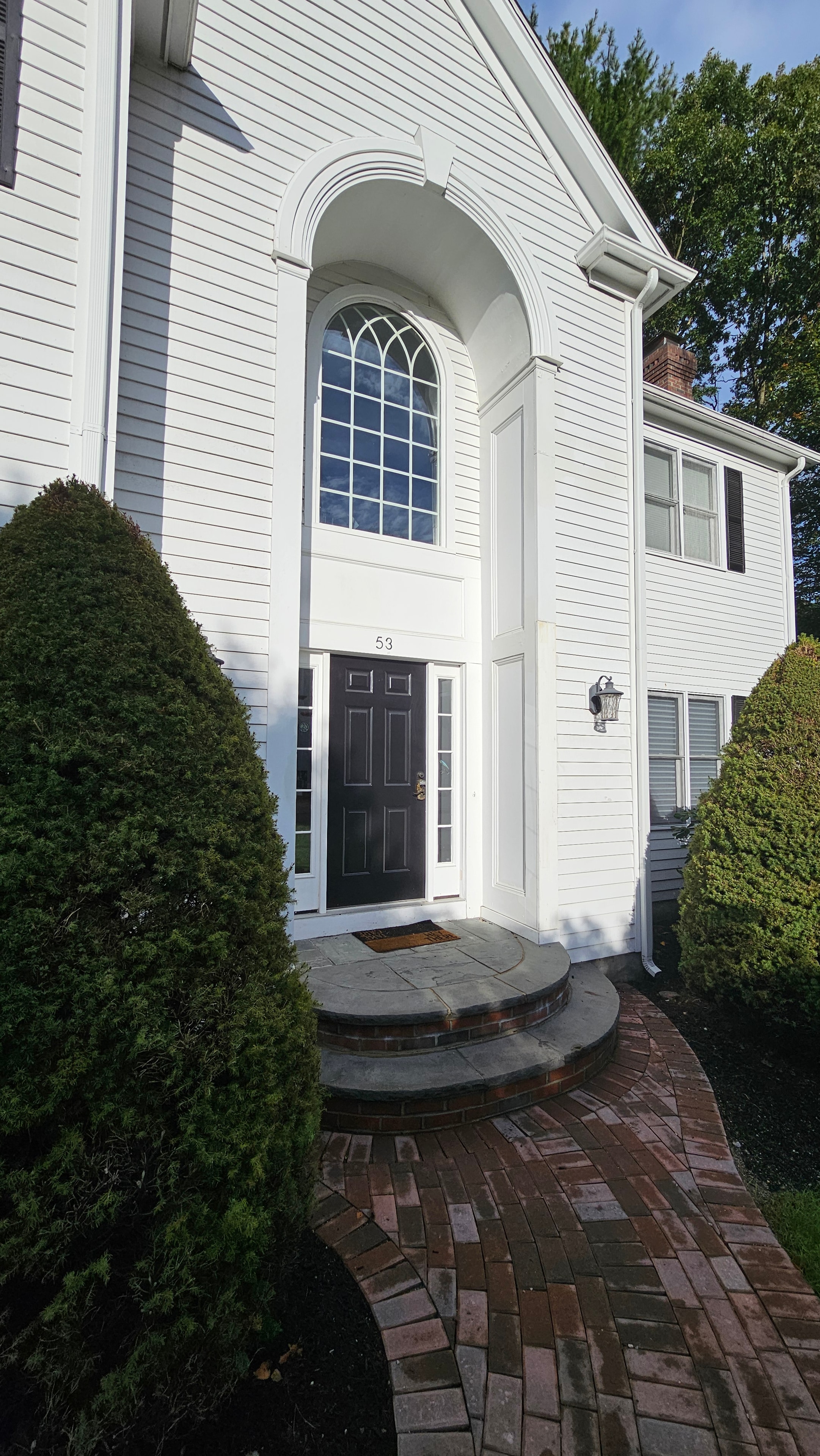 Finished front elevation of the Natick property after the multi-year restoration — white board-and-batten siding, dark front door, brick chimney, and arched entry.