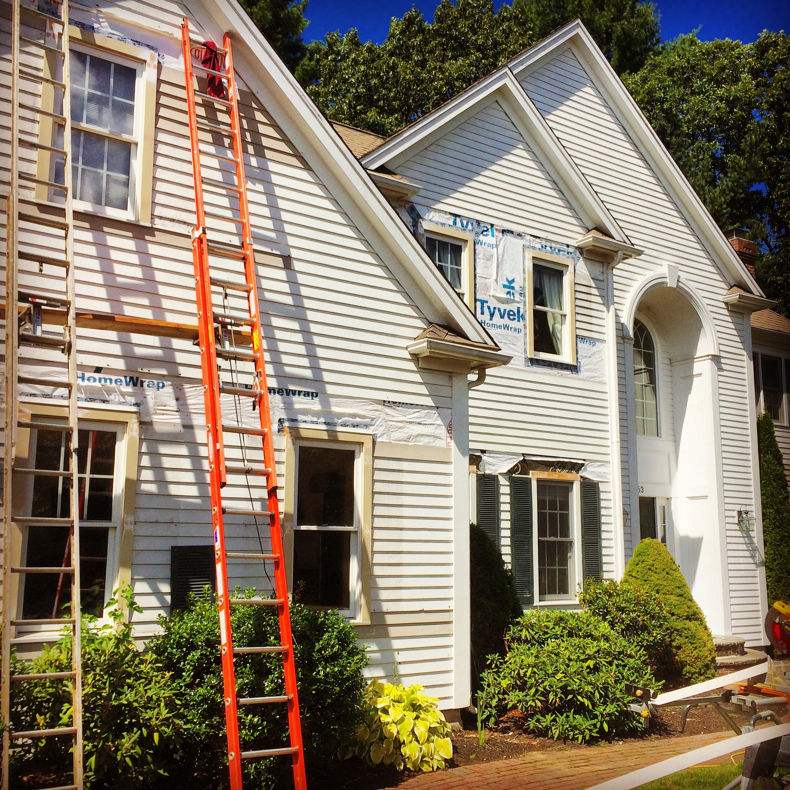 Front elevation of the Natick property during restoration, showing Tyvek HomeWrap on the upper gable and an orange ladder against the house.